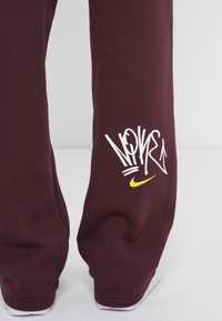 Bordeaux sweatpants lavet af blødt materiale; har hvid graffiti-lignende tekst og et lille gult Nike swoosh logo nederst.