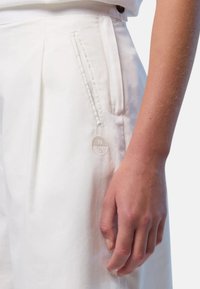 I pantaloni bianchi presentano tasche frontali e pieghe visibili. Il tessuto sembra liscio e leggero, con una sottile lucentezza. In primo piano si vede una mano appoggiata sul tessuto.