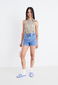 Grå ärmlös topp, högmidjade blå denimshorts, vita ankelsockor, blåa slides med logotyp, håller en liten vit handväska. Minimalistisk bakgrund.