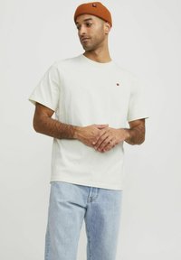 Krämfärgad bomullströja med korta ärmar, som har en liten röd pricklogga på bröstet och är matchad med ljusblå jeans.
