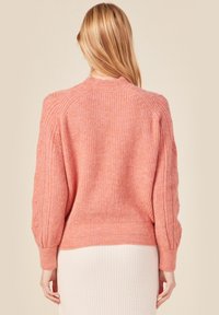 Rosa, stickad tröja med texturerad yta, har ribbad nederkant och puffärmar med knapprad längs armarna. Buren bakåtvänd.