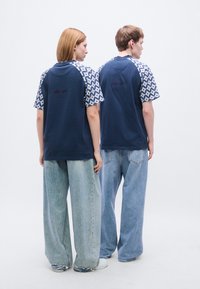 Μπλε ναυτικό μπλουζάκι με κοντά μανίκια με σχέδια, που φέρει ένα γραφικό "EST. 1970" στην πλάτη, συνδυασμένο με τζιν παντελόνι φαρδύ στο κάτω μέρος.