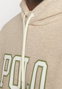 Béžová mikina z měkké tkaniny s velkým zeleným potiskem "POLO". Obsahuje bílé šňůrky na stažení a texturovanou kapuci.