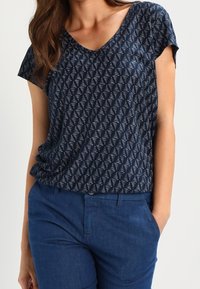 Marinblå V-ringad topp i lätt tyg med ett geometriskt triangelmönster; matchad med blå denimbyxor för en avslappnad stil.