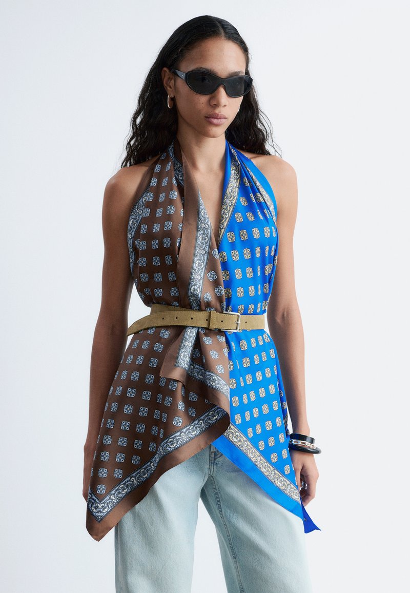 Vrouw met een zonnebril en een haltertop gemaakt van twee sjaals met patronen, blauw en bruin, met een ceintuur in de taille, en lichte spijkerbroek.