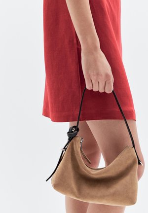 Sac en daim de couleur beige avec une forme courbée, une bandoulière en cuir noir et des détails zippés. Tenue par une personne portant une robe rouge.