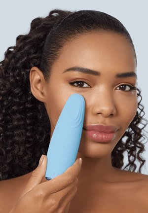 Femme aux cheveux bouclés tenant un appareil nettoyant pour le visage bleu clair contre sa joue droite sur un fond uni.