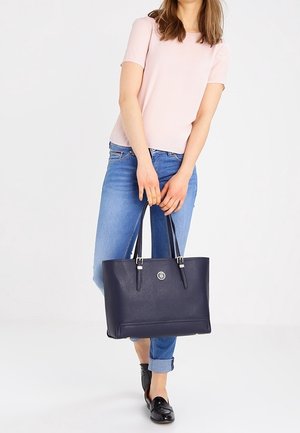 Marineblauwe shopper gemaakt van getextureerd materiaal met zilveren beslag, stevige handgrepen en een logo-accent. Draagt bij een roze top en spijkerbroek.