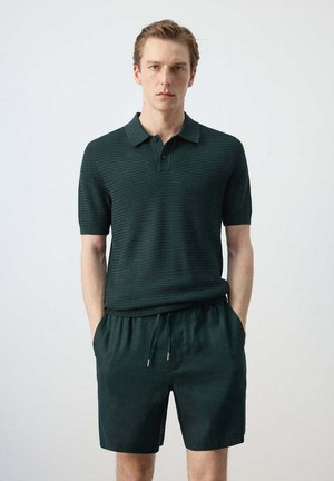 Homme debout, mains dans les poches, portant un polo à manches courtes texturé vert foncé et un short assorti sur un fond uni.