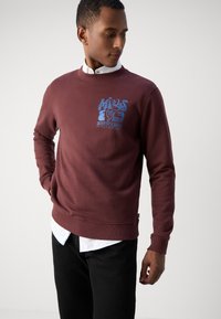 Felpa marsala con collo rotondo, maniche lunghe e design testuale grafico blu sulla parte anteriore. Realizzata in tessuto morbido e testurizzato.
