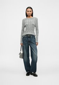Grauer Zopfmuster-Pullover, blaue weit geschnittene Jeans, schwarze Schuhe und eine gemusterte Tasche. Saubere Linien und lässige Passform mit strukturiertem Strickdetail.