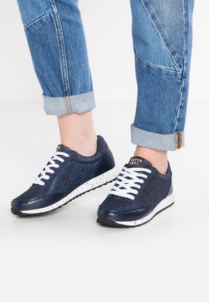 Stopy w granatowych, błyszczących trampkach z białymi sznurowadłami i białymi, nakrapianymi podeszwami, w połączeniu z podwiniętymi niebieskimi dżinsami na jednolitym tle.