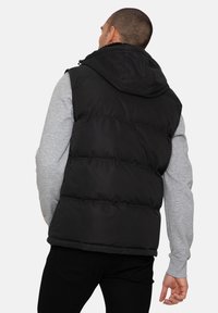 Schwarze gepolsterte Weste mit Kapuze, die eine glatte Außenseite, gerippte Säume und sichtbare Nähte aufweist. Trägt man über einem grauen Langarmshirt.