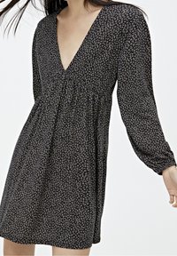 Donna che indossa un abito nero floreale con scollatura a V, maniche lunghe e vita arricciata, mostrata dalla gola fino a metà coscia su uno sfondo bianco.