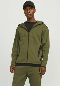 Sweat à capuche vert olive avec fermeture éclair sur le devant, accents noirs, deux poches latérales et une capuche structurée, porté avec une chemise noire en dessous.