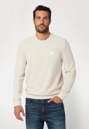 Cremfarbener Sweatshirt mit langen Ärmeln, Rundhalsausschnitt und weißem Logo-Patch auf der Brust, kombiniert mit dunkelblauen Jeans.