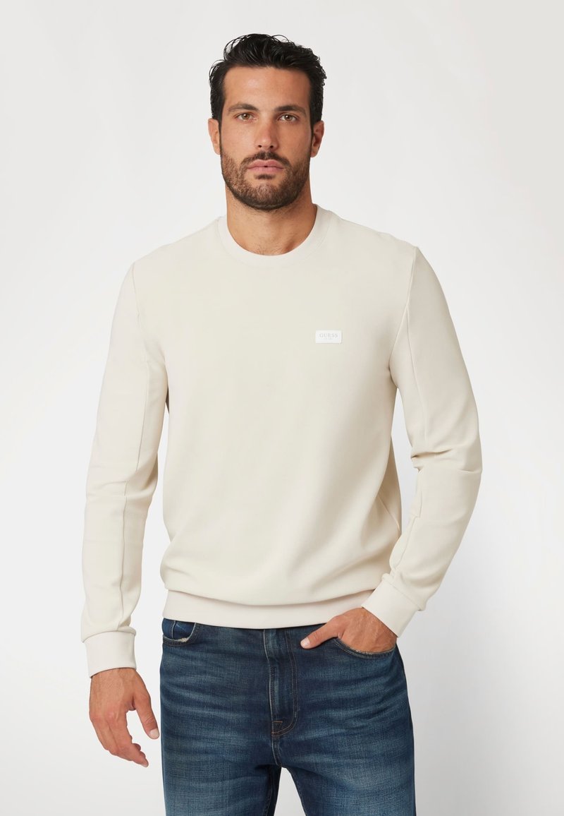 Krämfärgad sweatshirt med långa ärmar, rund halsringning och vit logotyp på bröstet, tillsammans med mörkblå jeans.