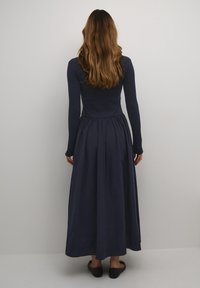 Long, navy blue dress made of soft fabric. Fitted bodice with long sleeves and gathered skirt. Worn with flat, textured black shoes.