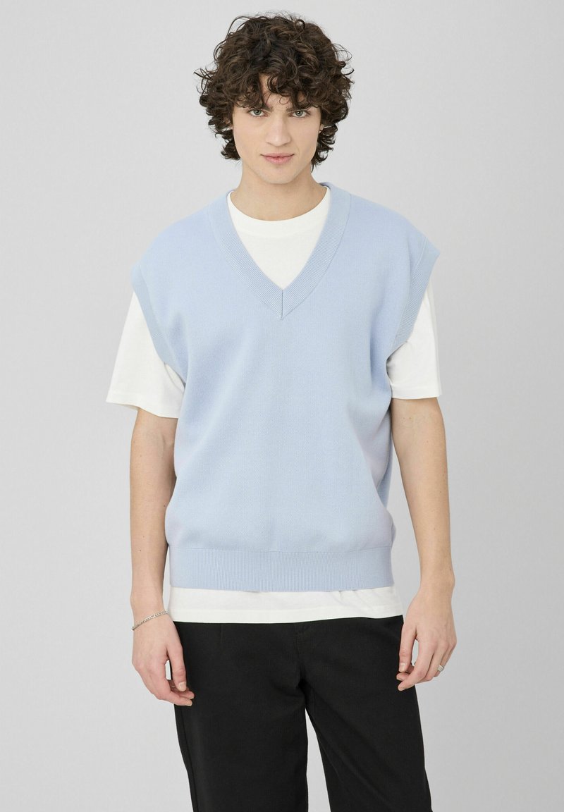 Veste sans manches en tricot bleu clair avec col en V, ourlet côtelé et texture, portée sur une chemise blanche. Le modèle se tient devant un fond neutre.