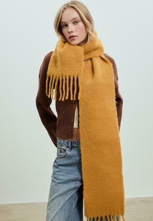Junge Frau, die einen dicken senfgelben Schal, eine kastanienbraune Kapuzenjacke und blaue Jeans trägt, steht vor einem neutralen hellen Hintergrund.