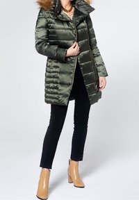 Abrigo acolchado verde oliva con capucha desmontable de piel, cierre delantero con cremallera y patrón acolchado diagonal. Combinado con pantalones negros y botines beige hasta el tobillo.