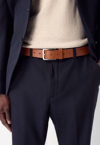 Ceinture en cuir marron avec boucle en argent, portée sur un pull crème et un pantalon bleu marine foncé, présentant une texture lisse et un design minimaliste.
