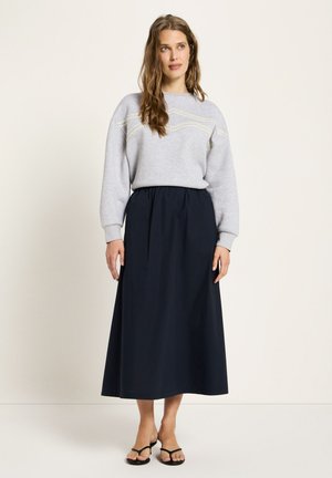 Woman wearing a light grey sweater with subtle yellow stitching and a high-waisted navy midi skirt, standing against a plain background.