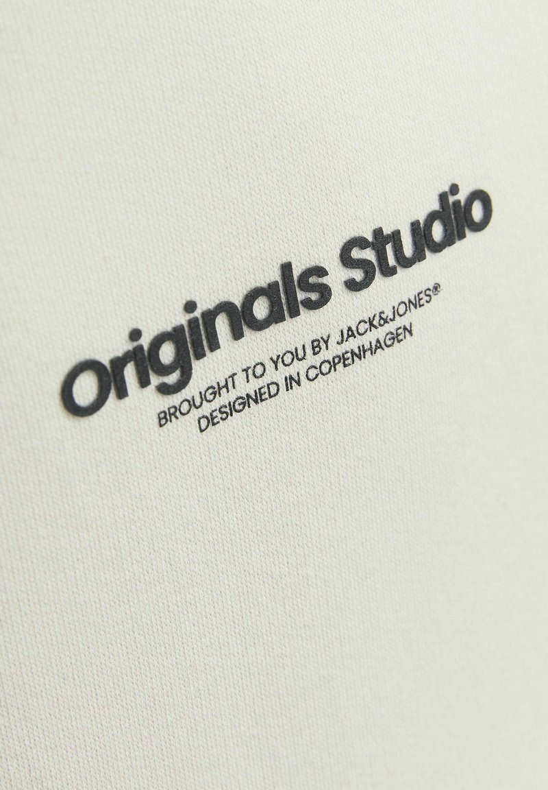 Tissu couleur crème avec une surface texturée, présentant le texte "Originals Studio" en noir gras, et une plus petite description en dessous.