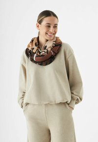 Écharpe infinie en tissu marron, beige et tan, avec une texture lisse et des motifs abstraits, portée sur un sweat beige clair.