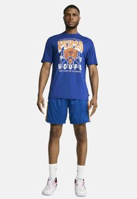 Kék Puma póló narancssárga grafikus puma logóval, kék csíkos rövidnadrággal, narancssárga oldalsó részletekkel párosítva. Fehér sportcipő és zokni.