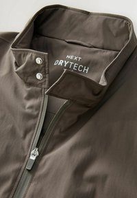 Veste marron foncé avec fermeture éclair et boutons-pression, avec l’étiquette "NEXT DRYTECH" imprimée à l’intérieur de la zone du col.