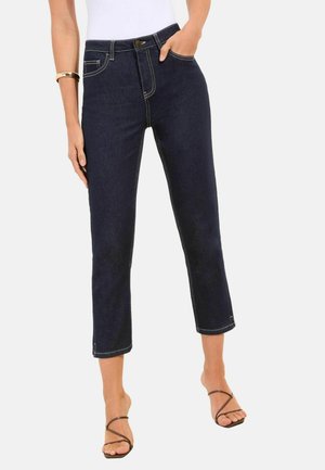 Žena nosiaca tmavomodré cropped džínsové nohavice s rovnou nohavicou a kontrastným prešívaním, biely top, zlatý náramok a čierne sandále na remienky, stojí s jednou rukou vrecku.