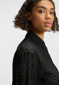 Camicia nera trasparente con colletto e dettagli con bottoni, caratterizzata da una sottile trama a motivi in una tonalità più chiara.