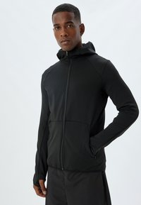 Giacca nera con zip e cappuccio, realizzata in tessuto liscio, con design aderente, aperture per il pollice nelle maniche e tasche frontali.