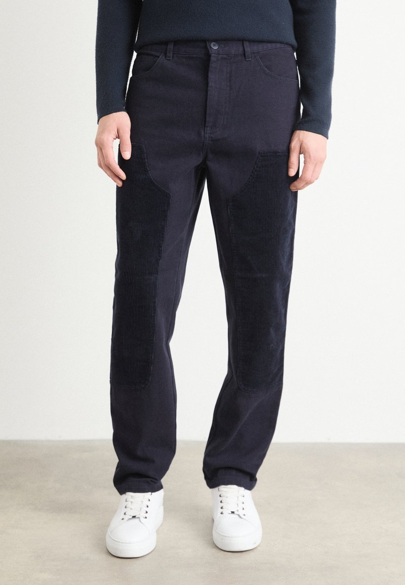 Marineblauwe corduroybroeken met textuurlapaccenten, rechte pasvorm, vijf-pocketontwerp en een knoopsluiting. Gecombineerd met witte sneakers.
