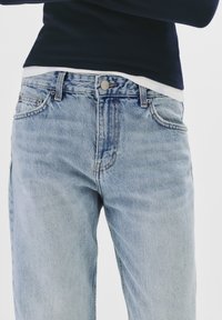 Los jeans de mezclilla de color azul claro cuentan con un diseño clásico de cinco bolsillos, con cierre de cremallera y botón. La tela presenta un ligero desvanecimiento y marcas de desgaste.