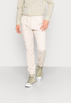 Mann trägt beige Cargohosen mit Kordelzug in der Taille und hellgrüne High-Top-Sneaker, steht vor weißem, einfarbigem Hintergrund.