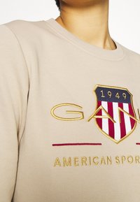 Sweat-shirt beige avec un écusson brodé. Présente des rayures rouges et blanches, des accents dorés, et un col rond côtelé.