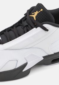 Białe trampki z czarnymi akcentami, z tekstylną cholewką, fakturowanym czarnym językiem i złotym logo Jumpman z boku.