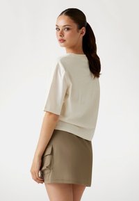 Crème top met korte mouwen en een subtiel reliëfpatroon, gecombineerd met een kaki mini-jurk met een grote zijzak. Eenvoudige texturen zijn duidelijk aanwezig.