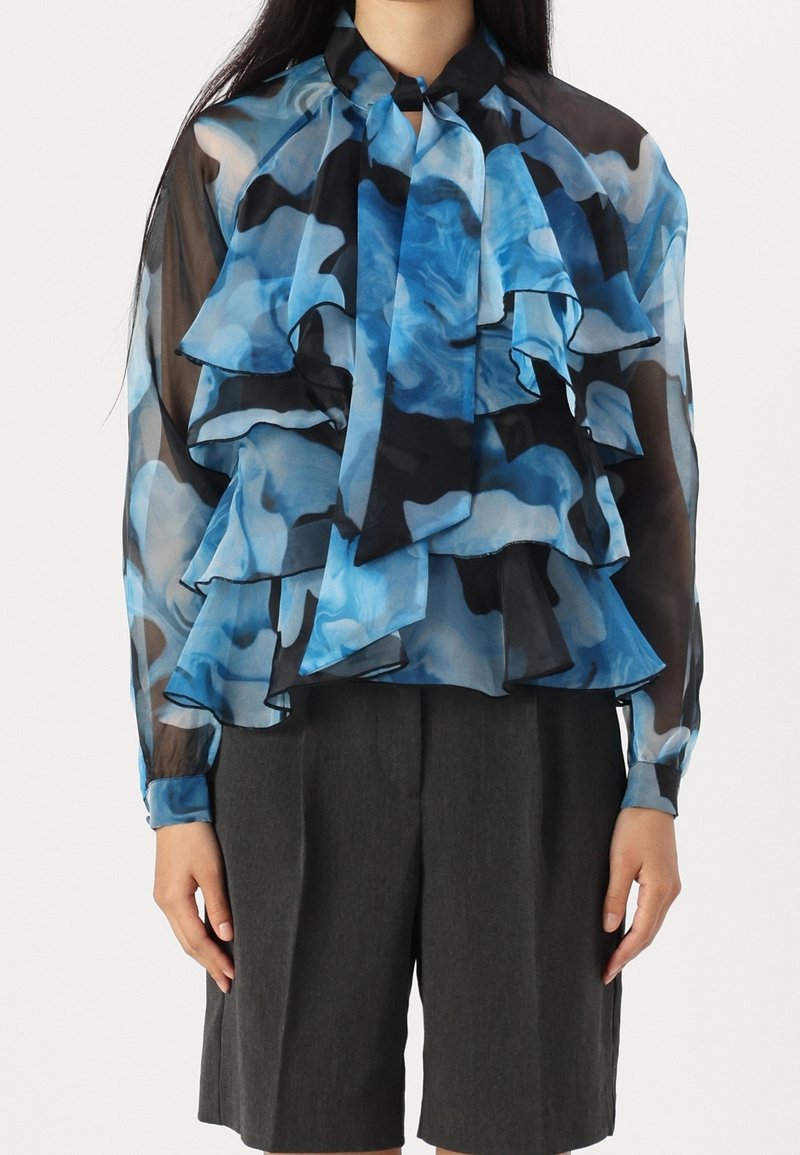 Blouse à motifs bleus et noirs en tissu transparent, avec des volants superposés, un col noué et des manches longues. Associée à un short gris.