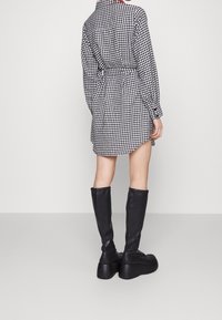 Vestido de camisa a cuadros en blanco y negro con cintura ceñida; mangas largas y collar. Llevado con botas de plataforma de cuero negro hasta la rodilla.