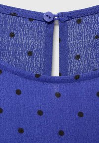 Blue fabric with a textured finish, featuring black polka dots and a button closure at the back. Smooth edges and a round neckline.