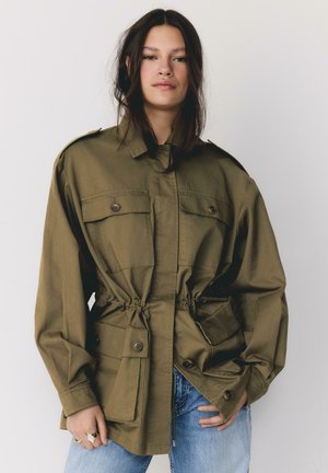 Mladá žena s dlhými tmavými vlasmi má na sebe nadrozmernú olivovo zelenú bundu s veľkými prednými vreckami a modré džínsy, stojí na jednoliatom pozadí.