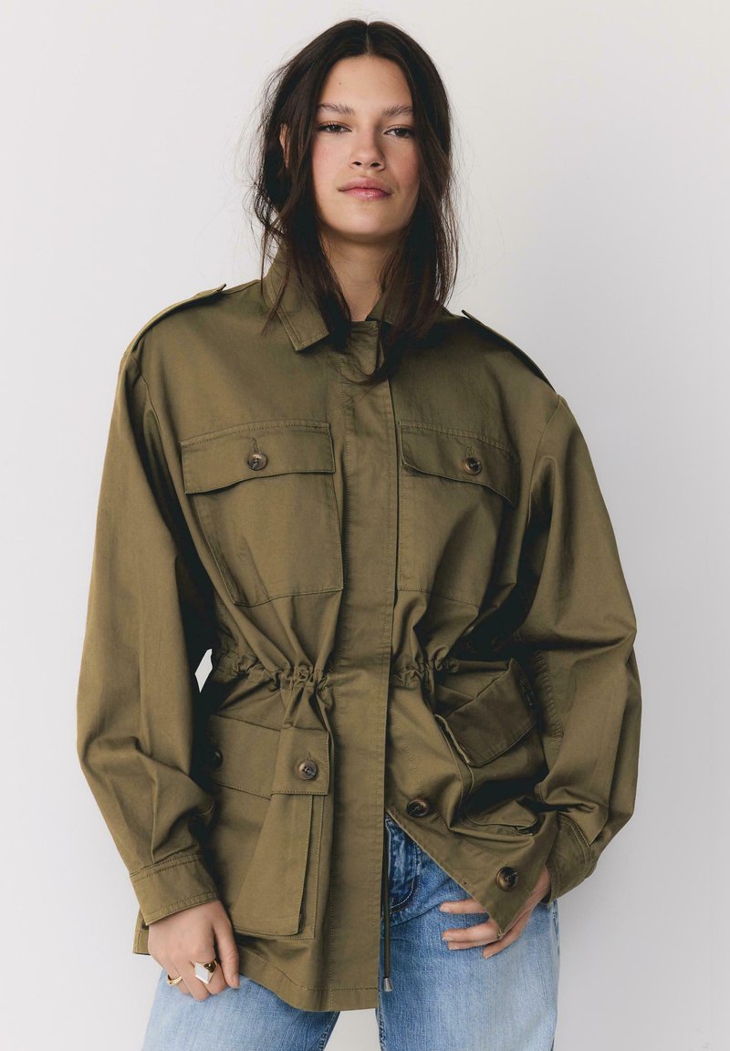 Jeune femme aux longs cheveux noirs portant une veste surdimensionnée vert olive avec de grandes poches avant et un jean bleu, debout devant un fond uni.