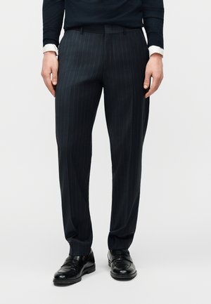 Man wearing black pinstripe dress pants, black loafers, and a dark sweater with a white shirt cuff visible.