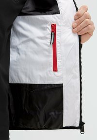 Interior de la chaqueta aislante con un forro de nailon blanco, un bolsillo con cremallera roja y una sección inferior negra. Textura duradera y suave.