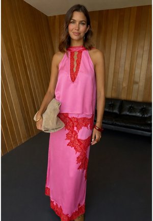 Femeie purtând o rochie halter din mătase roz cu detalii din dantelă roșie, ținând o poșetă bej, stând într-o încăpere cu pereți din lemn și o canapea din piele neagră.