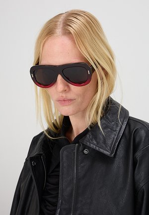 Donna bionda con occhiali da sole oversize con sfumatura dal nero al rosso e giacca in pelle nera con zip e bottoni a pressione.