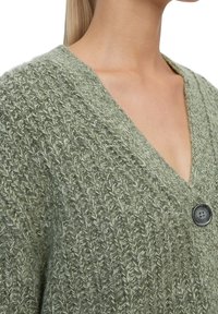 Grüne gestrickte Strickjacke mit V-Ausschnitt, strukturiertem Muster und einem einzelnen dunklen Knopf als Akzent. Der Stoff wirkt weich und etwas locker sitzend.